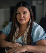 Brandi is wearing a blue shirt and sits with her arms on a table in front of her