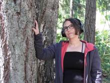 Sharon leans against a tree wearing headphones