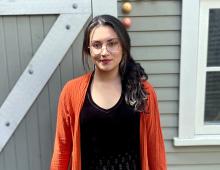 Sara Guzman stands in front of a building, wearing an orange sweater.