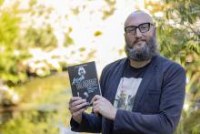 Dr. Theo Finigan, a VIU English Professor, holds up the book Atomic Blonde while standing in front of the VIU koi pond.